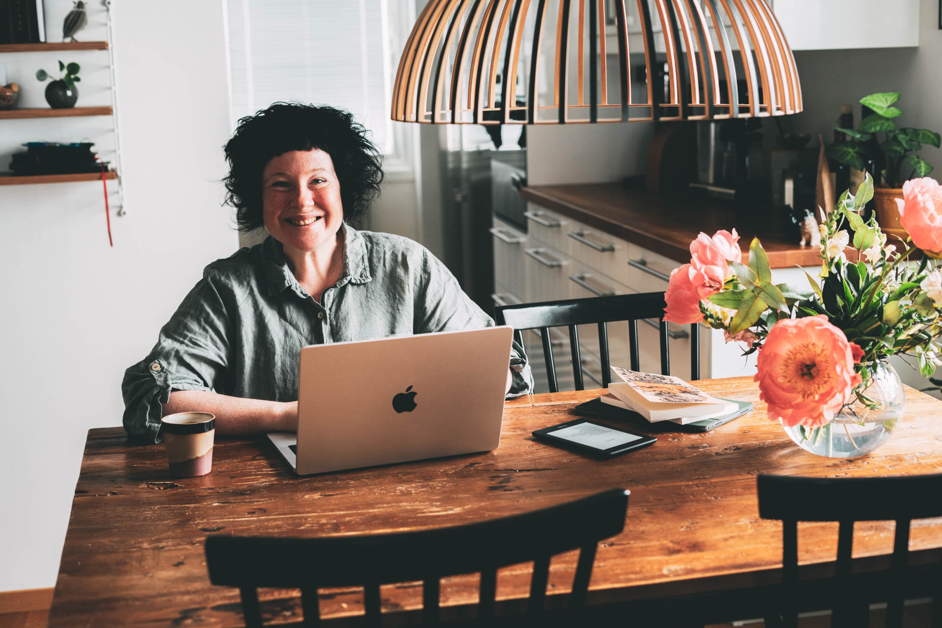 A smiling woman sitting at a kitchen table, working on a laptop.
